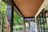 Wood ceiling panels installed on a covered porch, shown alongside dark wooden posts, a stone wall, black-framed windows, and outdoor greenery.